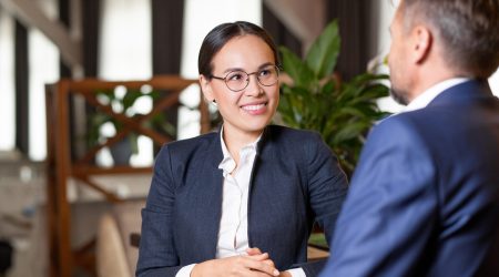 Pretty young cheerful agent in formalwear and eyeglasses looking at business partner or client during working discussion